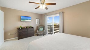 A bedroom with a bed, a ceiling fan, a dresser with decorative items, a chair, a TV, and a window with curtains. - Gulf View 10 by Liquid Life (Gulf Shores)