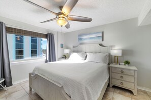 A bedroom with a bed, nightstands, a ceiling fan, a window with curtains, and a painting on the wall. - Royal Palms 1208 by Liquid Life (Gulf Shores)