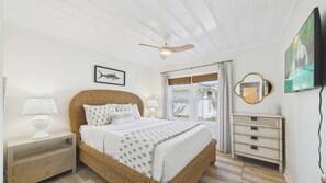 A bedroom with a wicker headboard, a bed with white bedding, a ceiling fan, a bedside lamp, a dresser, a mirror, and a TV.