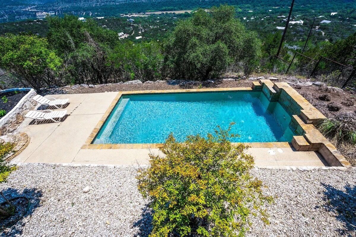 A rectangular swimming pool with clear blue water, surrounded by a stone border and a gravel area with lounge chairs.