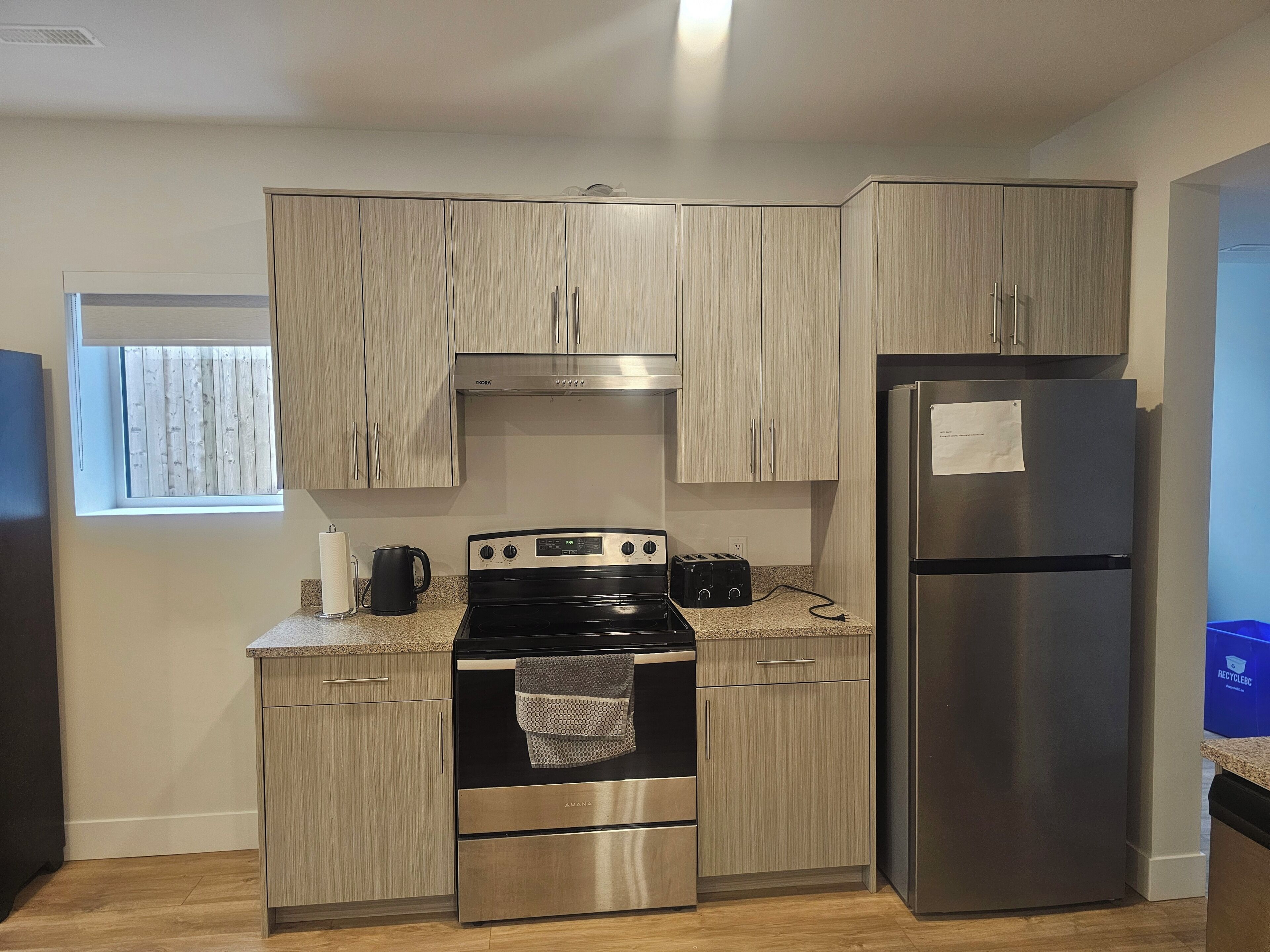 A modern kitchen with light wood cabinets, stainless steel appliances, and a window with blinds.