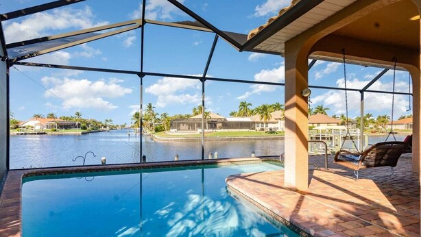 A pool with a view of a body of water and palm trees, a swing chair, and a covered area with a roof. - Rose Lake Escape - Gulf Access & Pet Friendly (Cape Coral)
