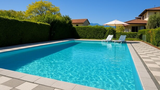 Ein rechteckiger Swimmingpool mit klarem, blauem Wasser, umgeben von einer gefliesten Liegefläche. Auf der rechten Seite befinden sich zwei Liegestühle mit Sonnenschirmen.