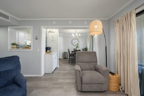 Un salon moderne avec un canapé gris, un fauteuil bleu et un autre fauteuil gris. On y trouve également un coin repas agrémenté d’un lustre et d’une horloge murale.