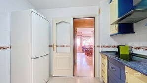 Una cocina con armarios blancos, un refrigerador y un comedor visible a través de una puerta abierta.