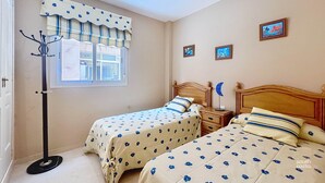 Un dormitorio con dos camas, una mesita de noche, una lámpara, un perchero y una ventana con cortinas.