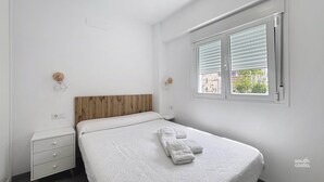 A bedroom with a wooden headboard, a single bed with a white bedspread, a nightstand with drawers, a wall-mounted light fixture, and a window with blinds. - Modern Central Apartment - South Costa, Nerja (Nerja)