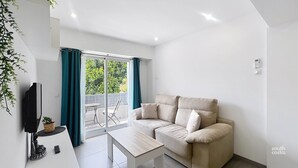 A modern living room with a beige sofa, a white coffee table, and a TV on a stand. There is a sliding glass door leading to a balcony with a view of greenery. - Modern Central Apartment - South Costa, Nerja (Nerja)