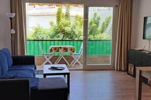 A living room with a sofa, a small table, a TV, and a balcony with a green fence and trees. - The Perfect Vacation in Nerja Near the Beach   (Nerja)