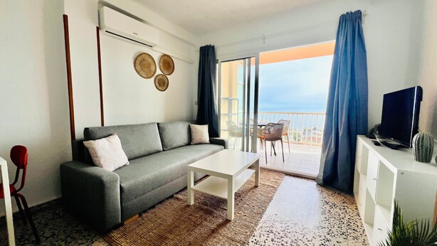 Una sala de estar moderna con un sofá gris, una mesa de centro blanca y un televisor sobre un mueble blanco. Se ve un balcón con cortinas azules y vistas al mar.