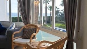 Habitación con vista a palmeras y una masa de agua a través de amplias ventanas. Hay una silla de mimbre con cojín y una mesita.