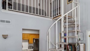 Ein geräumiger Wohnbereich mit hoher Decke, einer Wendeltreppe, einem Flachbildfernseher und einer durch einen offenen Durchgang sichtbaren Küche.