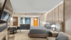 A modern hotel room with a bed, bedside table, lamp, and a seating area with a sofa and chairs.