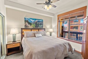Quarto com uma cama grande, duas luminárias de cabeceira, um ventilador de teto e um quadro de uma paisagem montanhosa.
