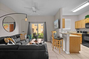 Un salon moderne avec un canapé, une table basse et un coin repas avec des chaises. On y trouve une cuisine dotée de meubles en bois et d’un four à micro-ondes. Un ventilateur de plafond est visible, et la pièce est éclairée par une grande fenêtre.