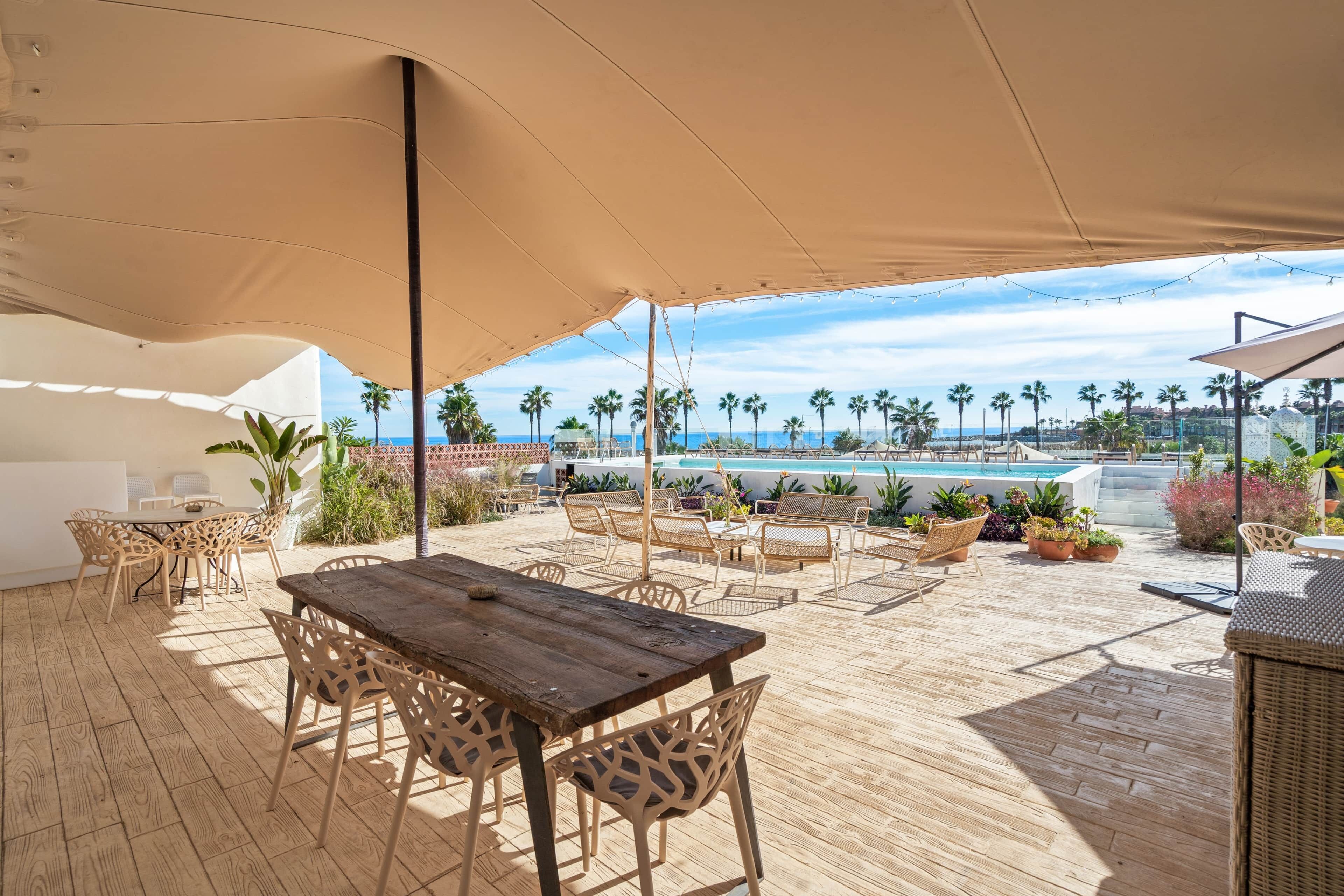 An outdoor patio with a wooden table and chairs, a pool in the background, and palm trees.