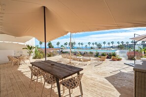 An outdoor patio with a wooden table and chairs, a pool in the background, and palm trees.