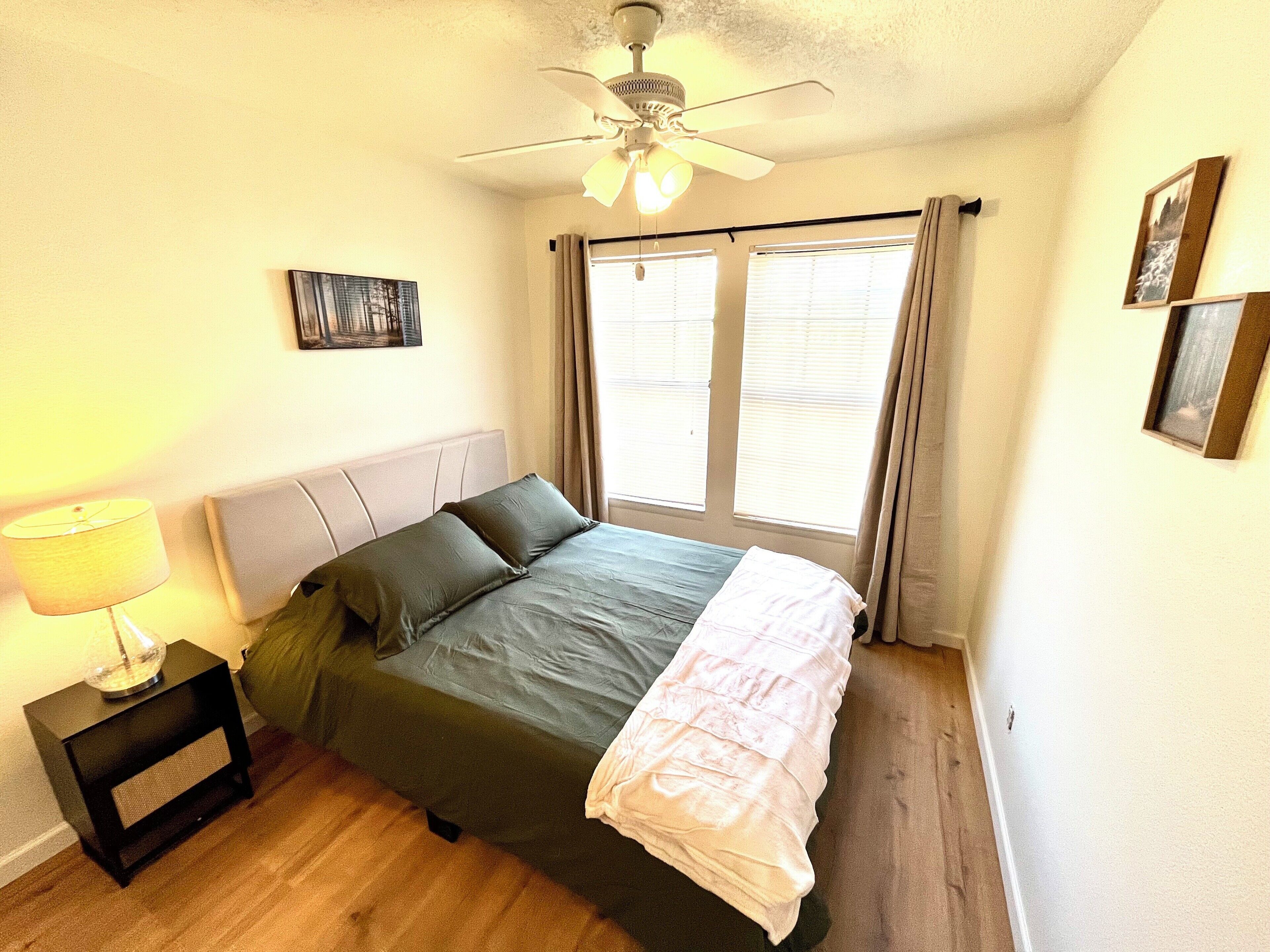 Une chambre à coucher comprenant un lit, une table de chevet, une lampe, un ventilateur de plafond et une fenêtre avec des rideaux.