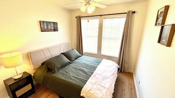 Une chambre à coucher comprenant un lit, une table de chevet, une lampe, un ventilateur de plafond et une fenêtre avec des rideaux.
