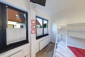 Ein Zimmer mit einem Etagenbett, zwei Fenstern mit Jalousien, einem Heizkörper und einem an der Wand befestigten Fernseher.