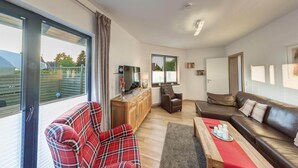 A modern living room with a sofa, armchair, and a dining table. There is a large window with a view of greenery outside. - Großzügige Ferienwohnung für bis zu 9 Personen (Burhave)
