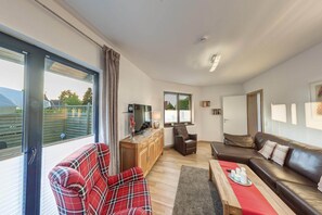 A modern living room with a sofa, armchair, and a dining table. There is a large window with a view of greenery outside.