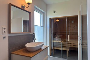 Moderni kylpyhuone, jossa on suuri peili, erillinen pesuallas ja lasiseinällä varustettu sauna.