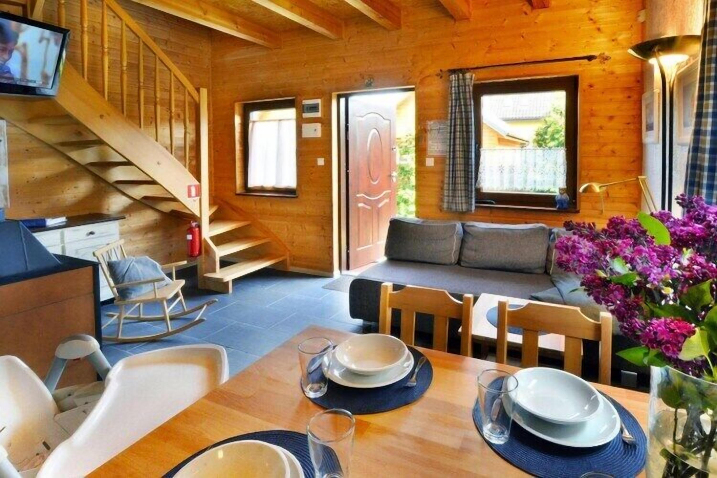 L’intérieur chaleureux d’un chalet avec un escalier en bois, un canapé, une table à manger dressée avec des assiettes et des verres, et un téléviseur fixé au mur.