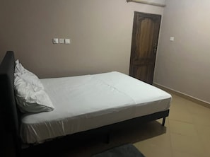 A single bed with a white mattress and a pillow, a wooden door, and a light switch on the wall. - 2-bedroom apartment in soothing Prampram with cool AC and gym access (Prampram)