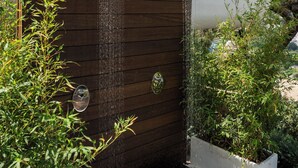 Una dutxa exterior moderna amb un fons de panell de fusta, envoltada de vegetació abundant i un cel blau clar.