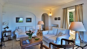Ein Wohnzimmer mit zwei Sofas, einem Couchtisch, einem Holzstuhl und einer Lampe. An den Wänden befinden sich Bücherregale und gerahmte Bilder.