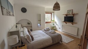 A bedroom with a bed, bedside table, lamp, TV, and a window with plants. - Lovely 3 bedroom detached house in Newbury (Newbury)