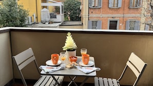 Un balcón con una mesa dispuesta para dos personas, sillas, una planta en maceta y un vaso de jugo de naranja.