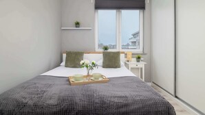 A modern bedroom with a large bed, grey bedding, a bedside table with a lamp, and a window with a view of buildings. - Modern Apartment | 2 Bedrooms | Workspace | Katowice. (Katowice)