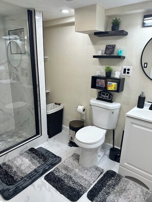 Een moderne badkamer met een douchecabine van glas, een toilet, een wastafel en een spiegel. Er zijn zwarte planken met toiletartikelen en een kleine plant.
