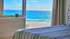 Une chambre dotée d’une grande fenêtre donnant sur une plage, d’un lit avec une literie à rayures et d’une petite table de chevet avec une horloge.