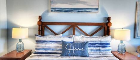 Una cama con una colcha a rayas, un almohadón con la palabra "Home" bordada y dos lámparas de noche.