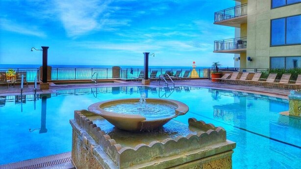 A swimming pool with a fountain, surrounded by lounge chairs and a view of the ocean. - Balcony | Walk to Pier Park | Beach Across Street (Panama City Beach)