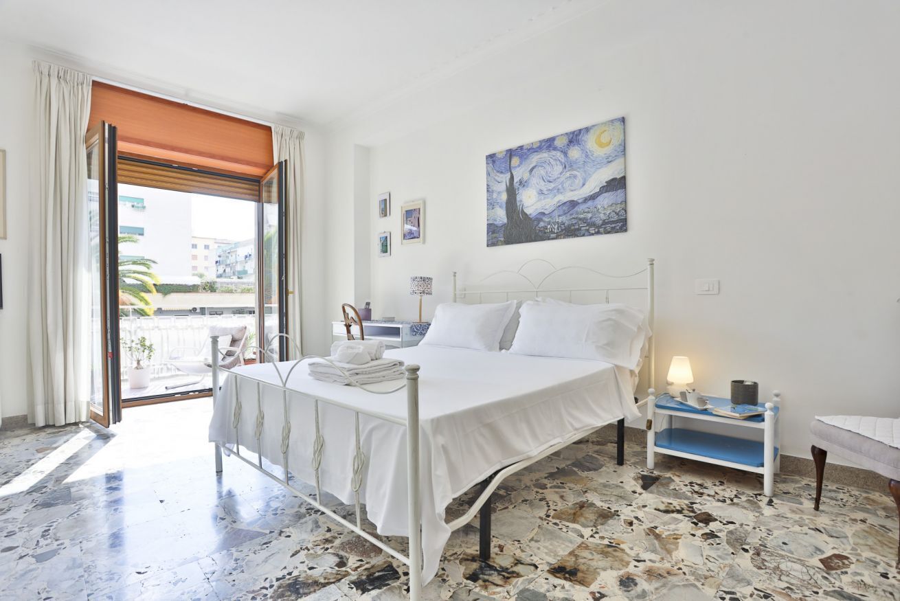 A bedroom with a bed, a nightstand, a painting, a window with curtains, and a view of buildings outside.