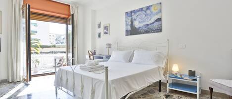 A bedroom with a bed, a nightstand, a painting, a window with curtains, and a view of buildings outside.