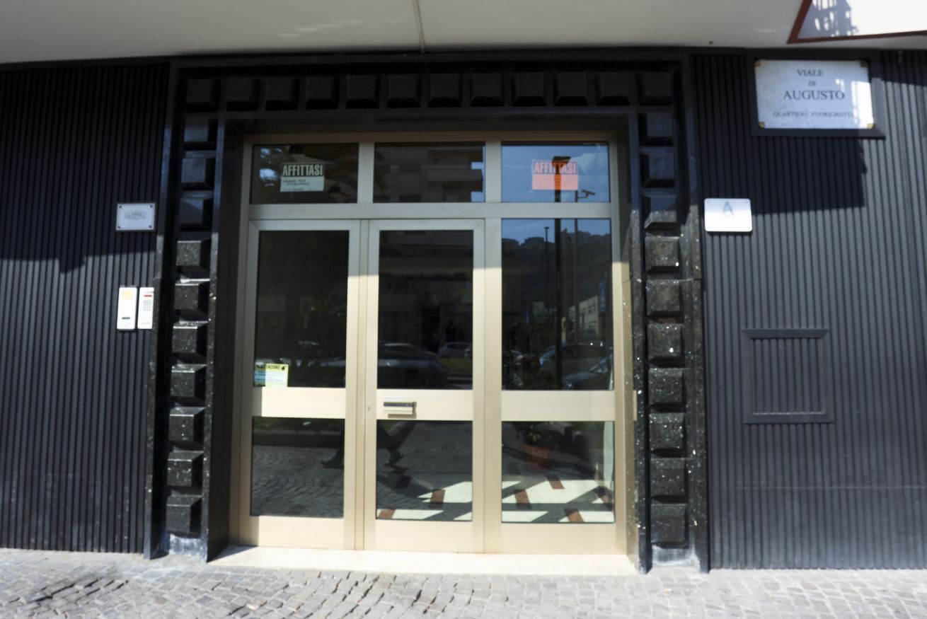 Ein Gebäudeeingang mit Glastür, einem Schild mit der Aufschrift 'AFFITTIST' und einem darüberliegenden Schild, das 'VIALE di AUGUSTO' ausweist.