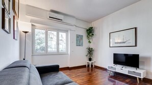 Ein modernes Wohnzimmer mit grauem Sofa, einem Flachbildfernseher auf einem weißen Standfuß, Holzboden, einem Fenster mit Jalousien und einem gerahmten Bild an der Wand.