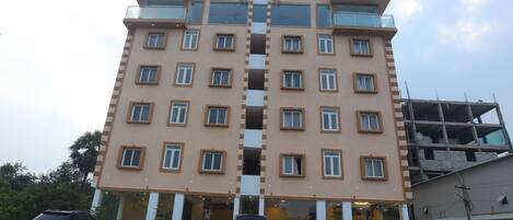 A multi-story hotel with a central vertical feature, balconies, and a parking area with vehicles.