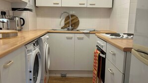 A compact kitchen with white cabinets, a wooden countertop, a sink with a wooden cutting board, a kettle, a washing machine, and a stove. - Stylish One Bedroom Apartment in Central Potton (Sandy)