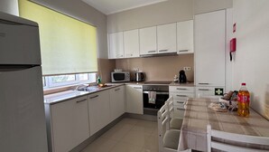 Eine kompakte Küche mit Kühlschrank, Mikrowelle, Herd und Spüle. Ein Esstisch mit Stühlen und ein Fenster mit Jalousien sind ebenfalls vorhanden.
