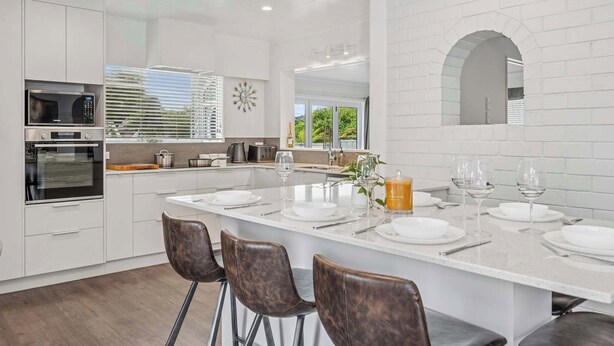 A modern kitchen with white cabinetry, a large island with bar stools, and a view of the outdoors through the window. - Tihi Time - Spa Pool, Games Room, Sleeps 10 (Rotorua)