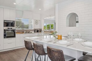 A modern kitchen with white cabinetry, a large island with bar stools, and a view of the outdoors through the window. - Tihi Time - Spa Pool, Games Room, Sleeps 10 (Rotorua)
