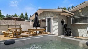 A pool area with wooden picnic tables and an umbrella, a hot tub, and a grill outside a building with a sloped roof. - Tihi Time - Spa Pool, Games Room, Sleeps 10 (Rotorua)