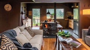 A cozy living room with a sofa, a coffee table, and a wood-burning stove. There is a window with a view of greenery outside. - Parque Cascadas, CABIN Rio Simpson-Patagonia Chile (Aysén)