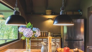 A kitchen with two pendant lights, a refrigerator, a wooden table with various kitchen items, and a window with a view of greenery. - Parque Cascadas, CABIN Rio Simpson-Patagonia Chile (Aysén)